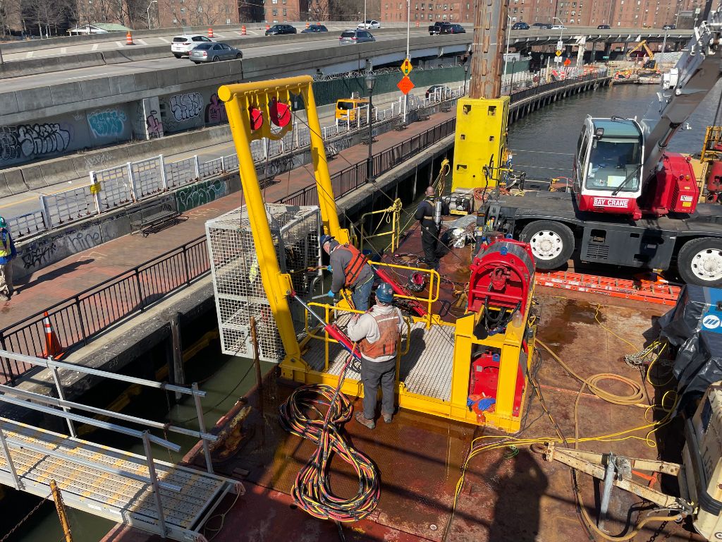 Overhead view of a full SubCon dive spread on a work barge — yellow LARS gantry, dive bell cage lowered into the water, dive supervisor and tenders managing umbilicals, Bay Crane in background, active urban waterway jobsite