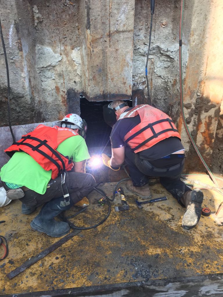 SubCon crew performing structural steel welding repair at a marine waterfront structure — two workers welding at the waterline