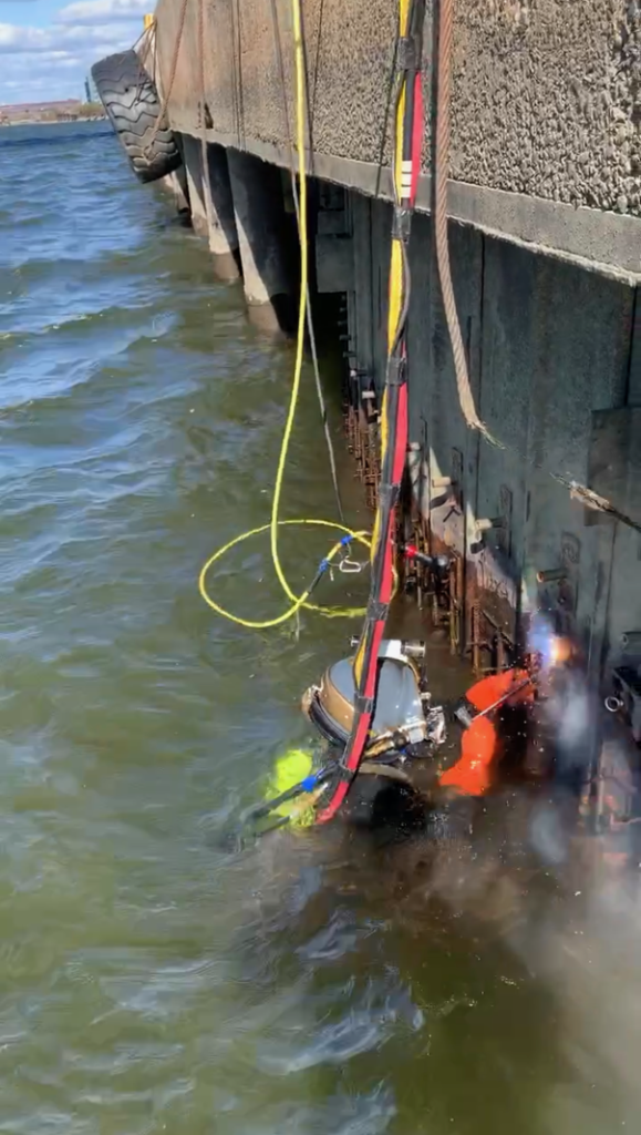 SubCon commercial diver performing underwater welding on submerged structural steel at waterline